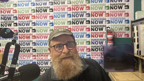 A bearded man sits at the desk with a microphone to one side of him. He is smiling and behind him the wall is covered in Subscribathon material. 