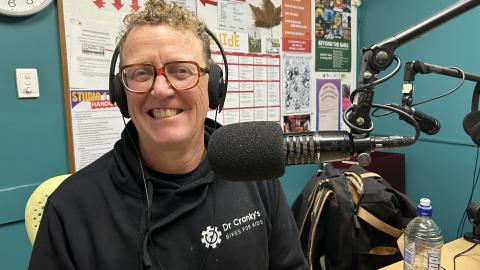 A smiling man wearing headphones sits next to a microphone in the 3CR studio. he is wearing a black sweatshirt with "Dr Cranky's Bikes for kids" written on it. 