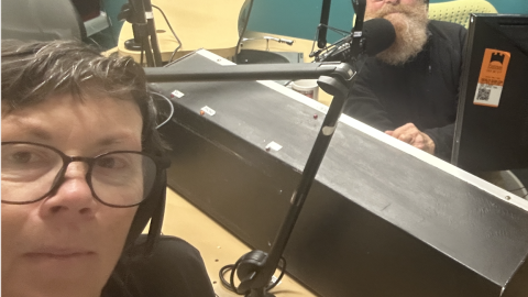 A woman wearing glasses looks into the camera, nehind her is a panelling dek and behind that a bearded man looking at the camera. 