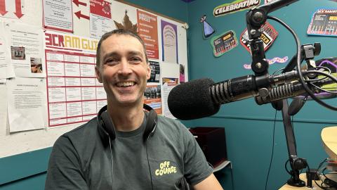 A smiling man in a green t-shirt with the logo "Off Course" on it sits next to a microphone and looks at the camera. 