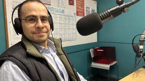 A man wearing headphones sits in front of a microphone and faces the camera