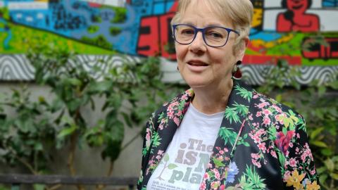 An older woman wearing a colourful jacket and short hair looks at the camera, she's standing outside in front of a mural and she looks content