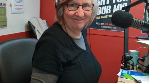 A woman with grey hair is looking at the camera, she is in a radio studio wearing headphones, glasses and a black shirt with a grey undershirt. 