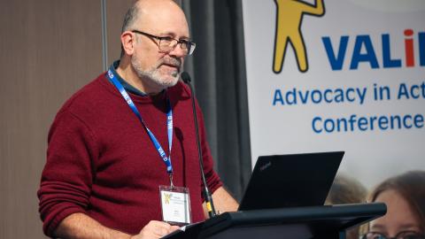 VALID CEO Fionn Skiotis, is standing at a podium and speaking into a microphone. He is wearing a red umper blue lanyard, and qlasses. Behind him is a white banner with vellow and blue graphics, text reads: 'VALID Advocacy in Action Conference'