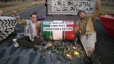 Jorgen Doyle and Yul Scarf sit on the access road to the Pine Gap military intelligence facility with a prop in colours of the Palestinian flag saying 'Close genocide bases, Land back, Liberation & Return'. Behind them, two people hold a banner saying, 'Stop the Genocide, Free Palestine, Close Pine Gap'.