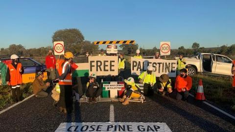 A photo of protesters blockading the Pine Gap access road in the early morning light. Two people are on the ground locked onto a barrel with a sign saying stop war crimes in Palestine, some are holding banners that say Free Palestine, and a large banner on the ground in front of the blockade reads Close Pine Gap Stop Genocide.