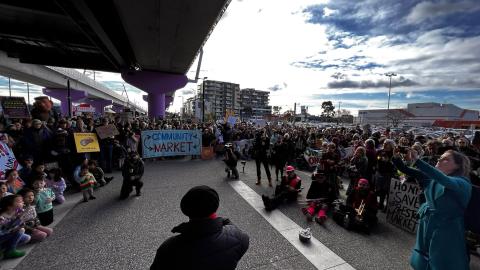 In the centre of the image we can see the back of Gaetano Greco as he addresses a gathering of people at the 2023 Hands Around Preston community protest to save the Preston Market from demolition and development. The gathering of people hold signs saying, 'Community Market,' and 'Honk to save our Preston Market'. 