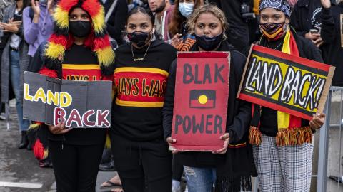 A photograph of four young Aboriginal women at the Invasion Day 2021 rally in Melbourne holding signs that read Land Back and Blak Power.