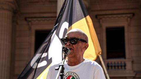 Professor Gary Foley speaking at the Naarm Invasion Day Rally 2026. He is wearing a shirt with a map of Palestine and the Camp Sovereignty flag flying behind him.