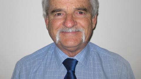 Neil Hunter is a caucasian man with grey hair and a moustache, wearing a powder blue collared shirt and a navy blue tie, he smiles at the camera against a white wall.
