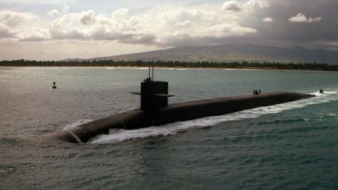Image of large black nuclear submarine in centre of photograph, travelling on surface of sea. There is a coast line behind and a mountain as well as a cloudy sky in background. 