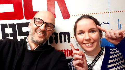 Photograph of presenters Brendan Bonsack (Left) and Carmen Main (Right), smiling at the camera. Behind them is a partial wall mural with the 3CR Radio logo.